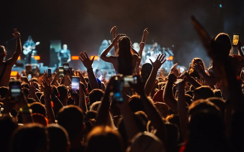 Young woman is sitting on someone's shoulders recording a concert on her smartphone while musicians are playing on stage