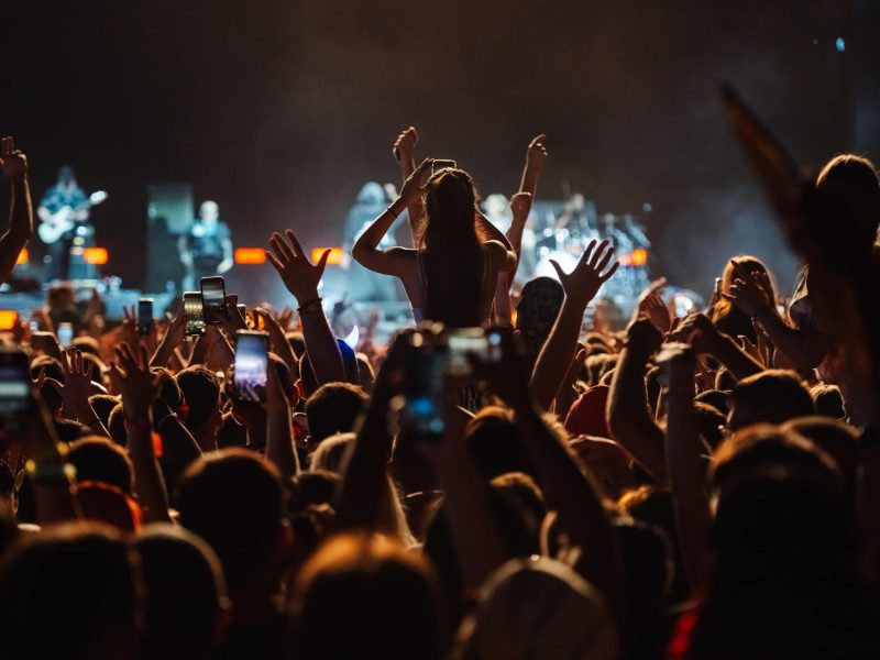 Young woman is sitting on someone's shoulders recording a concert on her smartphone while musicians are playing on stage